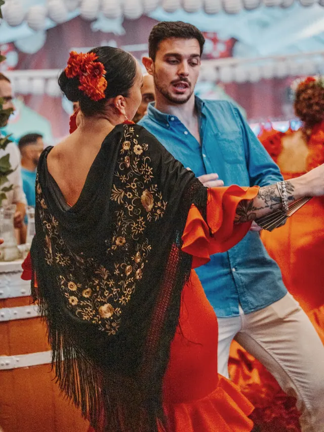 Pareja bailando en la caseta: ella, de espaldas, con mantón negro bordado y traje de flamenca naranja